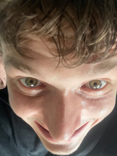 A close-up, low-angle shot shows the face of a person from the neck up, with their eyes looking directly at the camera. The individual has light brown hair and light-colored eyes with visible pupils and irises. Their nose is prominent, and their mouth is slightly open, revealing the inside of their mouth. The lighting creates highlights on their skin, particularly around the eyes and nose. The background is dark, suggesting an indoor setting. The image is a selfie taken from a low perspective. The specific location in the city of Brachterbeek, Netherlands, cannot be determined from the visual information provided in the image.
