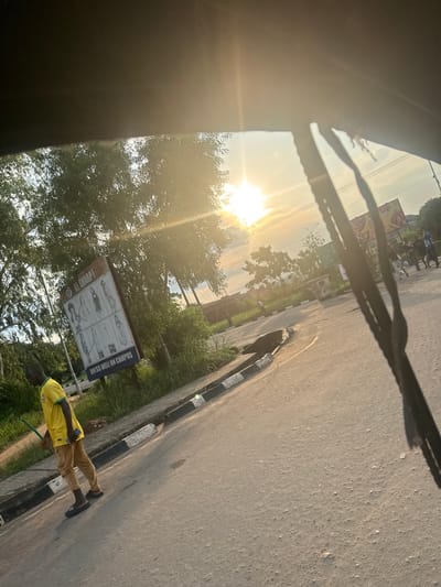 The image depicts a sunlit street scene in Amansee, Nigeria, likely during late afternoon or early evening due to the low angle of the sun casting long shadows and creating a golden hue. The camera appears to be positioned inside a vehicle, possibly looking out through a windshield, as indicated by the dark foreground elements that resemble a car's interior and possibly a wiper.
The scene includes a paved road with a distinct black and white striped curb. On the roadside, there is lush green vegetation and trees. A few people are visible: one individual in the foreground is wearing a bright yellow t-shirt and tan pants, holding a smartphone. Further down the road, a group of people are walking.
There is a sign on a pole by the roadside that shows several images of people and text. The text on the sign reads: "DRESS WELL ON CAMPUS!" and "TO BE SHARP!". This suggests the location might be near an educational institution or a commercial area that promotes a certain image. The overall atmosphere appears calm and ordinary, capturing a moment of everyday life in this Nigerian town.