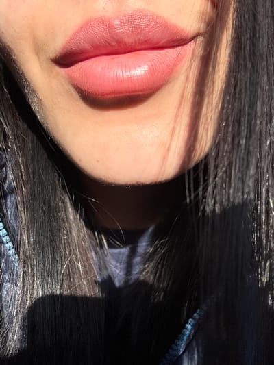 The image shows a close-up of a person's face, specifically focusing on the mouth and surrounding skin. The lips are painted with a pink lipstick, and the skin has visible pores and fine hairs. The hair is dark and appears to be lying around the face. The lighting is uneven, creating strong shadows on the face and hair. The presence of sequins on the clothing suggests a formal setting, and the scene location is Molodezhnyi, Russia.