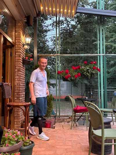 This image depicts an outdoor patio area in Cukarica Urban Municipality, Serbia, illuminated by artificial light.
One adult male is present, standing slightly left of the image center, facing partially right. He appears to be Caucasian, approximately in his 40s or 50s, with short, light brown hair. He is wearing a light blue short-sleeved collared shirt, dark blue shorts with a red vertical stripe on the outer seam, black compression socks extending to just below the knee, and multicolored athletic shoes (predominantly white, purple, and light green). His eyes are partially closed, suggesting a smile or squint.
The patio floor is composed of terracotta-colored paving tiles. To the far left, a brick column abuts a wooden doorframe and partially open door, suggesting an entrance to an indoor establishment. A dark-colored lantern with a warm glow is mounted on the brick column, with a small white security camera positioned directly below it. Two dark-colored planters containing pink/red flowers are attached to the brick column. A tall, dark wood bar stool with a woven seat and a small round wooden table are situated to the left of the male figure, near the brick column. In the bottom left foreground, a large dark planter contains green foliage and pink flowers.
The background is defined by a tall, dark green metal fence with a square grid pattern, supported by vertical green metal posts. Dense green trees are visible behind the fence. Two black hanging baskets with vibrant red geraniums are suspended from the metal fence. A small, conical evergreen shrub is planted on the ground directly behind the male figure, next to the fence. A red plastic watering can is visible on the ground behind the male's right foot, near the fence.
Multiple light olive-green metal chairs with perforated backs are arranged on the patio. One chair, positioned in the mid-ground, has a folded red garment on its seat. Two round, dark-colored metal tables are visible in the right half of the image; one has an ashtray. A dark wooden barrel serves as a base for one of the chairs on the far right. Overhead, a light-colored slatted awning or canopy is equipped with multiple embedded warm-toned lights, casting a glow on the immediate area.