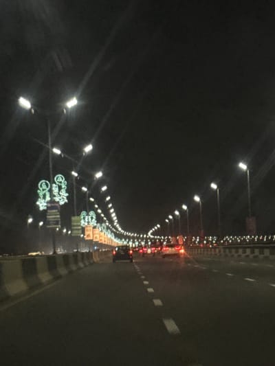 An aerial view of a dark, illuminated road. Lines of streetlights extend into the distance on the left, with decorative green lighting elements appearing below them. Traffic lights are visible on the right edge of the road. White pavement markings and striped dividers line the road's edge. Overall, the scene captures a night view of a highway or major road.
