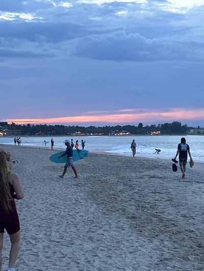 This is a vertical, low-angle shot of a beach scene at dusk in Arugam Bay, Sri Lanka. The sky displays a gradient of blues and purples, with streaks of orange and pink from the setting sun. The ocean meets the sandy shore, with small waves rolling in.
Several people are scattered across the beach and in the shallow water. In the foreground, a woman with blonde hair is lying on the sand, her back to the camera. Further up the beach, a person in a blue t-shirt and striped shorts is standing next to a surfboard. They appear to be preparing to paddle or are resting after a session. In the distance, a group of people are wading in the water or standing on the shore. One individual, wearing a dark t-shirt and patterned shorts, is holding two objects that resemble paddles or small surfboards, and another person is seen in the water nearby. A few more figures are visible further back, some appearing to be children or smaller individuals.
The overall atmosphere is relaxed and serene, suggesting an evening spent enjoying the beach and water activities. The time of day is clearly dusk, indicated by the colors in the sky and the dimming light. The presence of surfboards and paddles suggests surfing or paddleboarding is taking place, or has recently concluded. The background shows a treeline with some illuminated structures, hinting at a populated area or resort along the coast. No visible text is present in the image.