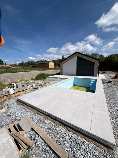 The image depicts an outdoor construction site in Horní Planá, Czechia, featuring a partially constructed swimming pool and surrounding patio area. The pool, filled with discolored water, is integrated into a light-colored tiled patio. To the right of the pool, blue steps are visible. The patio is edged with gravel and a metal drainage channel. In the background, a modern building with a wooden exterior and large dark doors stands adjacent to the pool area. Further back, a smaller wooden structure and a long stone wall with a black metal fence are visible. The site is littered with construction materials, including pallets, concrete blocks, and bags of material. The sky is blue with scattered white clouds, and the surrounding environment is rural, with trees and grassy areas. There are no people visible in the frame.