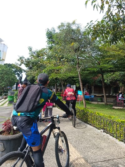 A man with a backpack is riding a bicycle on a path. He's wearing a colorful jersey and blue shorts. Another cyclist in a red jersey follows behind. The path is lined with a metal fence and greenery. People are scattered in the background, some seated at tables. Trees and a building are visible in the distance under a cloudy sky.