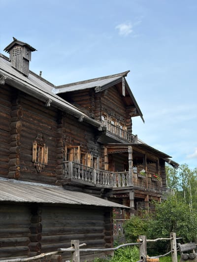 The image shows a wooden building with intricate carvings, typical of traditional Russian architecture, located in Nevskii Parkleskhoz, Russia. The building is constructed from dark, weathered logs, with several windows and balconies adorned with decorative wooden railings and trim. A prominent feature is a multi-story structure with a steeply pitched roof and a chimney capped with a wooden vent. The foreground shows a rustic wooden fence and greenery, suggesting an outdoor setting within a park or rural area. The sky above is partly cloudy and a light blue. There are no people or animals visible in the scene.