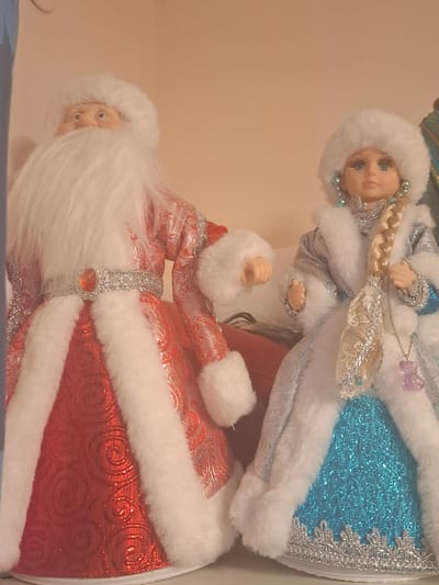 Two festive Christmas dolls, one resembling Santa Claus and the other a snow maiden, are positioned side-by-side against a plain, light-colored background. The Santa doll, on the left, wears a red coat with ornate silver patterns and white fur trim, a matching hat, and a prominent white beard. A silver belt with a large orange gem adorns its waist. The snow maiden doll, on the right, is dressed in a silver coat with white fur trim, a blue sparkly skirt, and a white hat. She has blue eyes, blonde braided hair, and wears large, ornate earrings. A silver chain with a small purple charm hangs around her neck. Both dolls are standing upright, with their hands visible. The image's lighting suggests an indoor setting, likely a shelf or display. The location in Mıñbaev, Kazakhstan, is not visually indicated within the image itself.