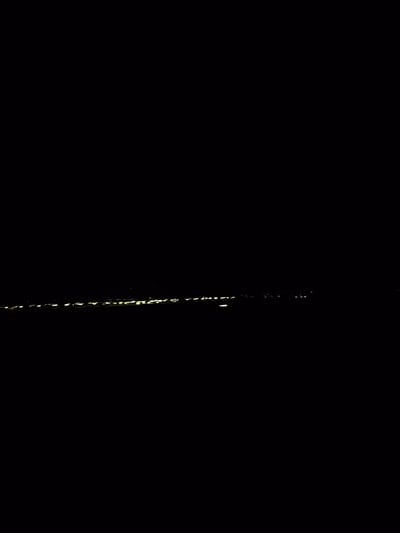The image shows a dark scene with a faint line of yellow lights on the horizon, suggesting distant city lights. The overall darkness implies it is nighttime. The context provided indicates the location is the Municipal Unit of Assiros, Greece. There are no visible people, objects, or actions. The environment is characterized by darkness and a distant, faint illumination.