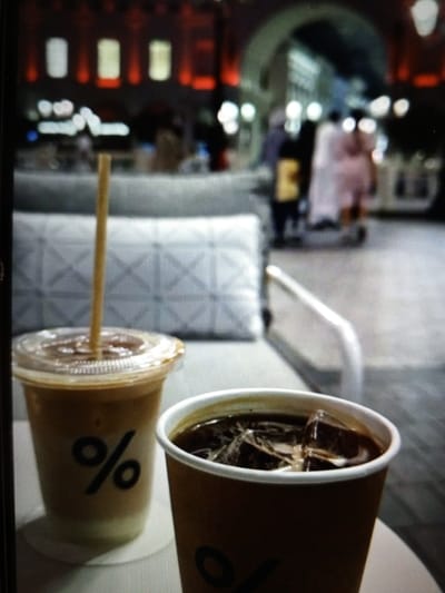 Two cold coffee drinks are placed on a table. The drink in the foreground is in a brown paper cup, filled with dark liquid and ice cubes. The second drink, to the left and slightly behind, is in a clear plastic cup with a clear lid and a brown straw. This drink has a lighter color, possibly iced coffee with milk or cream. Both cups have a black percentage sign logo. In the background, people are walking in what appears to be an outdoor market or plaza with a building featuring arched entrances and illuminated windows. The setting is a city street, potentially in Golbembal, Nigeria.