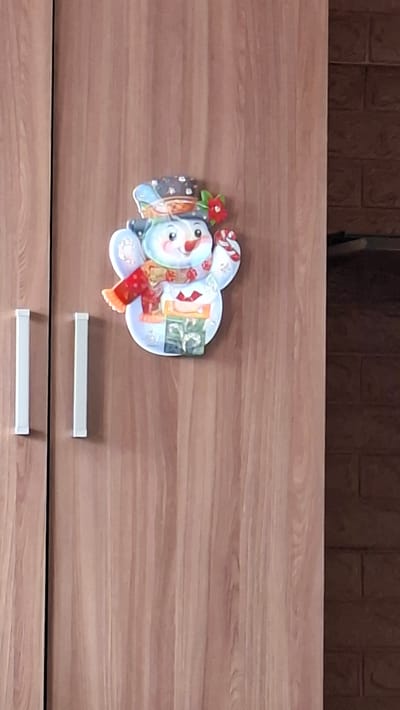 The image shows a close-up of a wooden closet door with a festive snowman decoration adhered to it. The snowman is cartoonish and appears to be made of thick paper or cardboard, with a blue hat, a red scarf, and holding a candy cane. It is positioned slightly off-center to the right. On the left side of the closet door, two white, rectangular handles are visible. The wood grain of the door is prominent. To the right of the closet door, a section of a dark brick wall is visible. There is no indication of the city of Novoilinsky, Russia, in the image.