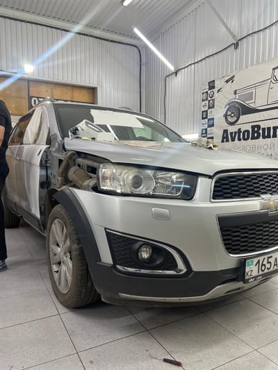 The image shows a silver Chevrolet Captiva parked in a garage. The car's hood is open, and some parts appear to be disassembled, suggesting it is undergoing repair or maintenance. The garage has a tiled floor and white metal walls, with fluorescent lighting illuminating the space. A banner with the text "AvtoBuren" and a graphic of a car is visible on the wall. The car's license plate, "KZ 165A", is also visible. A red-handled screwdriver lies on the floor near the front bumper, indicating active work is in progress. The scene appears to be in a professional auto repair shop. No specific time of day or weather conditions can be determined from the image.