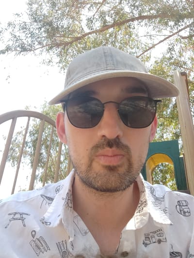 A man wearing a light-colored baseball cap, sunglasses, and a white short-sleeved shirt with a print of various icons including a picnic table, a map, diving fins, a camper van, and a grill is captured in a close-up, outdoor, selfie-style photograph. He has a goatee and is looking towards the camera. The background consists of trees with visible branches and foliage, a metal fence, and a colorful structure that appears to be part of a playground. The lighting suggests it is daytime. The scene is outdoors, possibly in a park or recreational area, within the city of Yoav Regional Council, Israel.