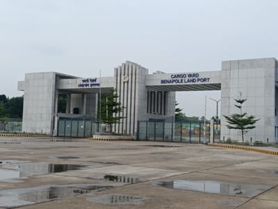 The image shows the entrance to a cargo yard at Benapole Land Port in Benapole, Bangladesh. The entrance is a large, modern concrete structure with the words "CARGO YARD BENAPOLE LAND PORT" written in blue letters above the entryway. To the left, in Bengali script, is text that appears to read "বেনাপোল স্থল বন্দর". The entrance features a central section with vertical fluted columns and a circular emblem. Large metal gates are visible behind the entrance, leading into the port. The ground in front of the entrance is paved concrete with several large puddles reflecting the sky and the structure. Small trees are planted around the entrance, and yellow and black striped barriers are present. The sky is overcast. No people or vehicles are clearly visible in the immediate foreground or at the entrance.