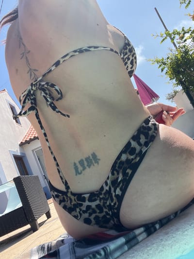 A close-up, waist-up shot of a person wearing a leopard-print bikini in an outdoor setting. The person is positioned with their back to the camera, and their skin is visible, along with a tattoo on their back. The background includes a bright blue sky, some greenery, a white building with red-tiled roofs, and outdoor furniture. The image appears to have been taken during the day, with bright sunlight casting shadows. The presence of a swimming pool and lounge chair suggests a leisure location.