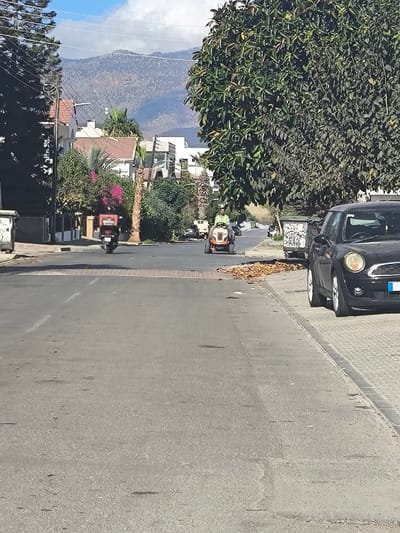 A residential street scene in Nicosia, Northern Cyprus / Cyprus, is depicted under clear daytime conditions with a partly cloudy sky and distant mountains. The asphalt road, marked with a dashed white center line, extends into the background. A reddish-brown paved speed bump crosses the road in the midground.
On the immediate right, a black Mini Cooper is parked on a light-colored paved sidewalk. Adjacent to the parked car, further down the sidewalk, is a graffiti-covered green public waste bin. A pile of dry leaves rests on the edge of the sidewalk and road near the bin. A large, dense green tree with broad leaves dominates the right side of the frame. On the immediate left, a dark-colored public waste bin and a tall, narrow pine tree are visible.
Two individuals are operating vehicles on the road, moving away from the viewer. On the right side of the road, a person wearing a light green shirt and dark trousers drives an orange ride-on lawnmower. Further ahead and to the left, another person on a black scooter or moped, equipped with a red insulated delivery box, also travels away. The scooter operator is wearing a dark helmet.
Residential buildings with light-colored facades, red-tiled roofs, and various trees including palm trees and flowering bougainvillea line both sides of the street. In the background, a range of arid, hazy blue-grey mountains is visible beneath the sky.