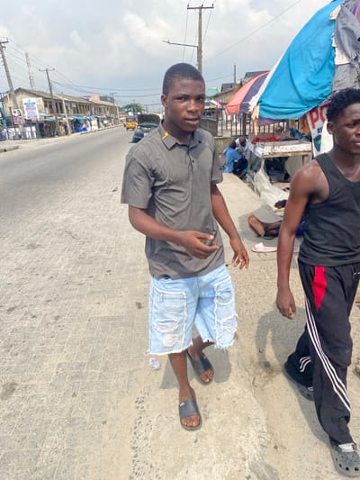 The image displays an outdoor urban street scene in Langbasa, Nigeria, under a bright, partly cloudy sky. In the immediate foreground, a young Black male stands, positioned slightly to the viewer's right and directly observing the camera. He wears a dark grey short-sleeved polo shirt with a yellow stripe on the collar, light blue distressed denim shorts with frayed hems, and dark grey slide sandals. His left arm is slightly bent, with the hand open. To his right, a second young Black male is partially visible, from the chest down, walking leftward. He is dressed in a black tank top, black track pants with red accents and three white stripes on the outer leg, and black Crocs-style footwear.

The ground beneath them is a paved sidewalk constructed from interlocking grey bricks, with scattered small pieces of debris. Adjacent to the sidewalk on the right, several makeshift market stalls are present. One prominent stall features a light blue tarp canopy and displays red produce, identified as tomatoes, in a tray. Several individuals are seen gathered around these stalls, with one pair of bare feet visible on the ground.

In the mid-ground, a paved asphalt road extends into the distance. A yellow tuk-tuk is visible on the road. On the left side of the road, a row of low-rise commercial buildings lines the street, some displaying signage such as "WELLCARE PHARMACY" and "AVON." Numerous utility poles with overhead electrical wires crisscross against the sky, indicating infrastructure.