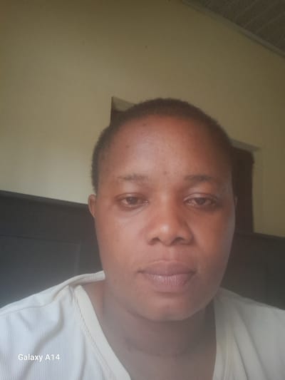 A close-up, slightly angled shot of a woman's face, possibly a selfie, taken indoors. The woman has short, dark hair and appears to be wearing a light-colored, ribbed top. The background consists of a dark, undefined area and a light yellow wall. The location, Udi, Nigeria, is not visually confirmed in the image. The woman is looking at the camera. Text "Galaxy A14" is visible in the upper left corner.