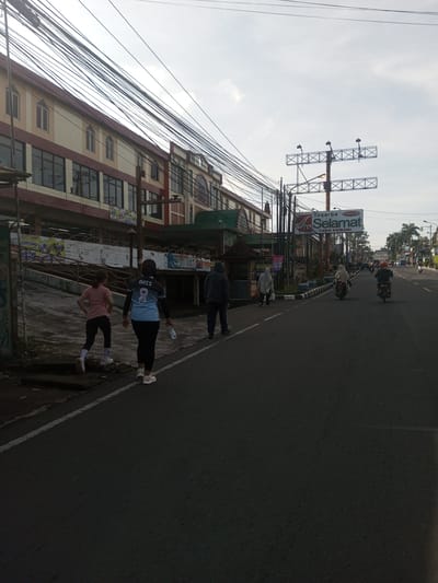 The image shows a street scene in what appears to be a town or city. A multi-story building with large windows and a beige and red facade is visible on the left side of the frame, partially obscured by a low wall or barrier covered in advertisements. Power lines are densely arranged overhead.
Several people are walking on the side of the road. Two women, one in a pink shirt and dark leggings and the other in a light blue sports jersey with the name "ANIS" and the number "8" on the back, and black leggings, are walking to the left. Behind them is a man in a dark hoodie, and further down the road is another person wearing a hijab.
Two motorbikes are travelling on the road. A sign advertising "Toserba SELAMAT" is visible above the road, attached to a metal structure. Further down the street, additional vehicles are visible. The sky is overcast.
There is no indication that the location is Phou Khoun, Laos.