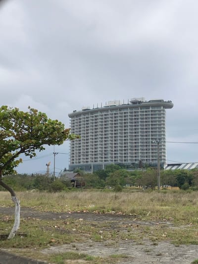 The image displays an urban and undeveloped landscape in Hải Vân Ward, Vietnam, under an overcast sky. A prominent, light-colored multi-story building, estimated to be 18 stories tall, dominates the background. This structure features a grid-patterned facade of balconies or windows and a distinctive flat rooftop with a dark, curved, cantilevered platform extending to the right. The foreground consists of an unmaintained, grassy field with patches of bare dirt and scattered debris. A tree with a white-painted lower trunk and green foliage is positioned in the left foreground. In the midground, a utility pole with overhead wires is visible, alongside various green trees and shrubs. A rustic, single-story wooden building with a dark, sloped roof is partially obscured by trees in the mid-left. A yellow construction crane is faintly visible in the distant left background. No individuals are present in the scene.