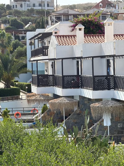 The image shows a multi-story white building with dark wooden balconies and a red-tiled roof in Chersonisos Municipal Unit, Greece. The building is surrounded by lush green vegetation, including palm trees, hedges, and prickly pear cacti. In the foreground, there are two thatched umbrellas and two orange life preservers visible near what appears to be a swimming pool or water feature. The background reveals more buildings on a hillside, also with red-tiled roofs. The sky is clear and blue.