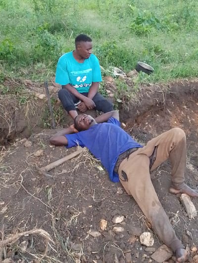 In Otulu, Nigeria, two individuals are pictured in an outdoor, rural setting. One man is seated with his legs crossed, wearing a turquoise t-shirt with a graphic design and dark pants. He appears to be looking off to his right. The second man is lying on his back on the ground, with his head resting on his hands. He is wearing a blue t-shirt and tan pants, one of which has a visible tear. A shovel lies on the ground near him. The scene is characterized by rough, disturbed earth, suggesting manual labor or excavation. In the background, green foliage and vegetation are visible, indicating an outdoor environment. A black pot is also present among the rocks and debris. The lighting suggests it is daytime, with no specific indication of weather conditions. The overall impression is one of rest or respite after some form of work.