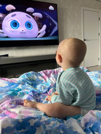 This full-shot indoor photograph, captured in Dubovka, Russia, shows a light-haired baby positioned on a colorful bed or blanket, engrossed in watching a television screen. The baby, visible from a rear-side perspective, is wearing a light blue and white striped long-sleeved top and a diaper. Their back of the head, left ear, and bare feet are discernible. They are lying face-down, with their head turned to the left, towards the screen.

The baby is situated on a vibrant blanket or duvet cover featuring an abstract floral pattern in shades of blue, purple, and pink. To the left, a flat-screen television displays an animated character. This character is light purple/white with large, bright blue eyes, small ears, and visible claw-like appendages. The text "ani" is partially visible in the top-left corner of the TV screen.

The background reveals a light-colored, textured wall. In the upper-right portion of the image, a white access panel or hatch, equipped with two black latches, is mounted on the wall, possibly leading to an attic or utility space. The room is brightly lit, suggesting it is daytime. The focus is clearly on the baby and the television program.