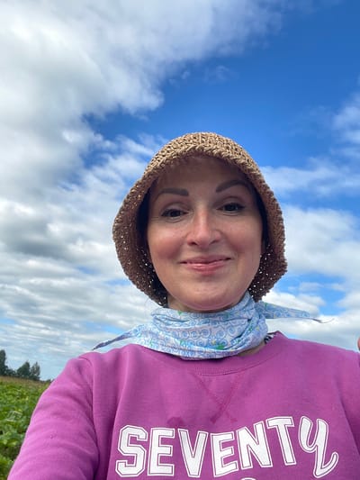 A medium shot captures a woman wearing a straw hat and a pink sweatshirt in an outdoor setting with a blue sky and clouds. She has brown hair and is smiling at the camera. The sweatshirt has the word "SEVENTY" printed on it in white letters. She is also wearing a light blue scarf with a white swirled pattern around her neck. In the background, there is a field of green plants and trees. The location is identified as Dresvyanovo, Russia.