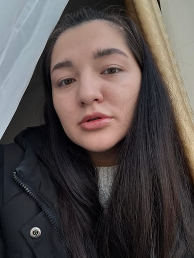 The image displays an upward-angled, medium close-up self-portrait of an adult female. She has long, dark brown hair falling over her shoulders, brown eyes, and full, dark eyebrows. Her lips are slightly parted and appear glossy. She wears a black zipped jacket with a silver snap button visible on the left chest area, over a light grey knitted sweater. The subject gazes directly into the camera. The background consists of a translucent white curtain on the left side and a textured, light brown curtain on the right, indicating an indoor setting adjacent to a window. The image provides no specific visual information to identify the location as Kurovskoye, Russia, or any external environment.