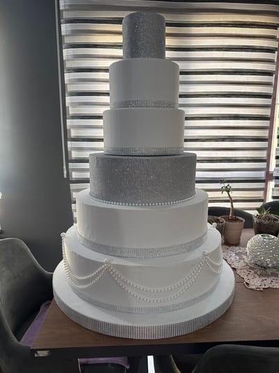 A five-tiered white and silver glitter cake stands on a wooden table. The cake is decorated with rhinestones and pearl strands. In the background, a striped blind is visible. The scene is indoors, with a chair visible on the left side of the image. The cake is the main subject.