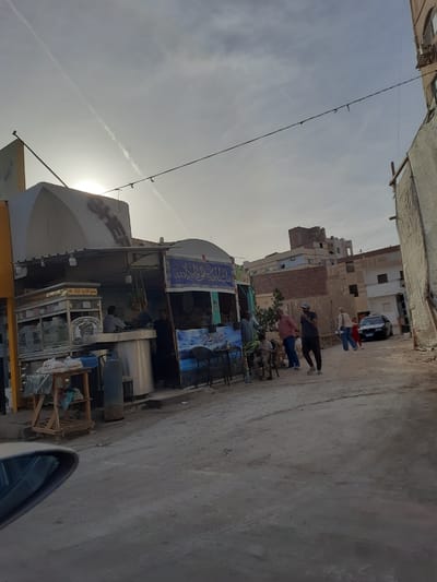 The image depicts a street scene in Hurghada, Egypt. Several people are standing and walking along the side of the road, near a row of small shops or stalls. The buildings in the background are typical of the architecture in the area. A dark-colored car is parked further down the road. There is a noticeable absence of vegetation. The sky is overcast, and the sun appears to be partially obscured by clouds. The scene gives the impression of a typical day in a somewhat busy, local area.