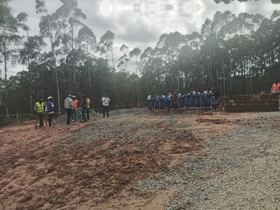 The image captures an outdoor gathering in Moticho ward, Kenya, against a backdrop of tall, slender trees under an overcast sky. The foreground consists of reddish-brown earth and piles of grey gravel, indicating a site under development.
On the left, a group of men are present, some wearing construction safety vests (yellow, blue, and orange) and hard hats. Two men are holding shovels, suggesting work-related activities. One person is actively recording the event with a mobile phone. Near the center, another individual in a light-colored shirt (possibly pink) is also holding a phone or recording device.
On the right side of the scene, a larger group of young people, mostly girls, are standing in multiple rows. They are uniformly dressed in blue garments, possibly school uniforms, with white tops. One person among them holds what appears to be a drum or a similar traditional instrument. Further to the right, a partially constructed low brick wall or foundation is visible, with another person in an orange safety vest standing nearby.
The overall impression is a community event, possibly a groundbreaking ceremony, an official visit, or an inspection related to a new construction project, involving both community members, students, and possibly construction personnel or officials. The weather is cloudy, and the time of day is daytime. No visible text is discernible.