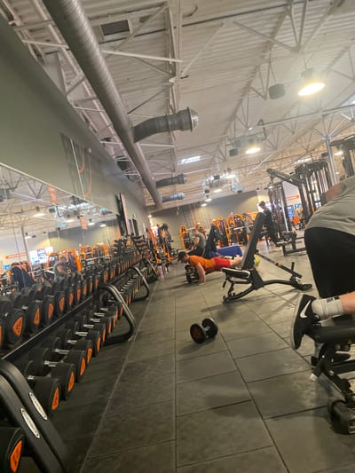 The image depicts the interior of a Basic-Fit fitness center located at 1 Rue Vasco de Gama, 14200 Hérouville-Saint-Clair, France. The scene is brightly lit by overhead industrial lighting and features a high ceiling with exposed metal trusses and silver corrugated air ducts. The floor is covered with dark gray interlocking rubber tiles.
In the foreground on the left, a long, low rack of black dumbbells with orange weight indicators extends into the background, receding in perspective. The dumbbells range in size and are arranged neatly. A large mirror runs above this dumbbell rack, reflecting parts of the gym.
In the mid-ground, a male individual wearing an orange t-shirt and red shorts is performing push-ups on the floor, utilizing two small black hexagonal dumbbells as handgrips. He is positioned next to a black adjustable utility bench. Two loose small black dumbbells are on the floor slightly closer to the camera.
To the immediate right in the foreground, a partial view of a person's lower body is visible, with legs bent and feet (wearing black Nike sneakers with white socks) resting on a black gym bench, obscuring the upper body.
Multiple other individuals are visible throughout the background, engaged in various exercises using weight machines which are predominantly black with orange accents, consistent with Basic-Fit branding. One person is seen using a leg press or similar machine further back. The gym appears well-maintained and active, with various pieces of equipment and several patrons utilizing the facility.