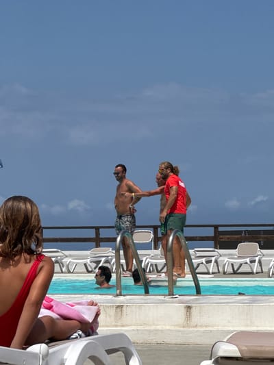 The image captures a sunny day scene at a poolside in Capaci, Italy. In the foreground, a woman with brown hair is seated on a white lounge chair, wearing a red swimsuit and holding a pink towel. To her right, two men stand by the pool. One man, wearing patterned swim trunks and sunglasses, gestures with his hand. The other man, in a red t-shirt with the word "LIFEGUARD" printed on it and green shorts, appears to be in conversation with the first man. Further into the pool, another man is partially submerged in the water. The background features a wooden railing, more lounge chairs, and a clear blue sky with scattered clouds. The pool itself has bright blue water.