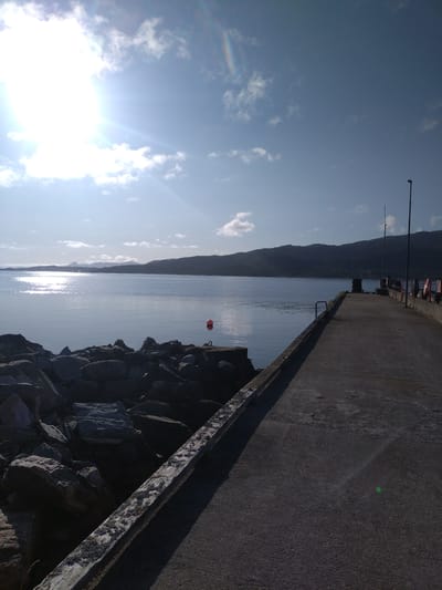 The image shows a concrete pier extending into a calm body of water, with a rocky shore to the left. Hills form the distant horizon, and the sky is blue with scattered clouds and bright sunlight. A small orange buoy is visible in the water. On the pier, there are lampposts and railings. The scene appears to be the harbor area of Bremsnes, Norway.