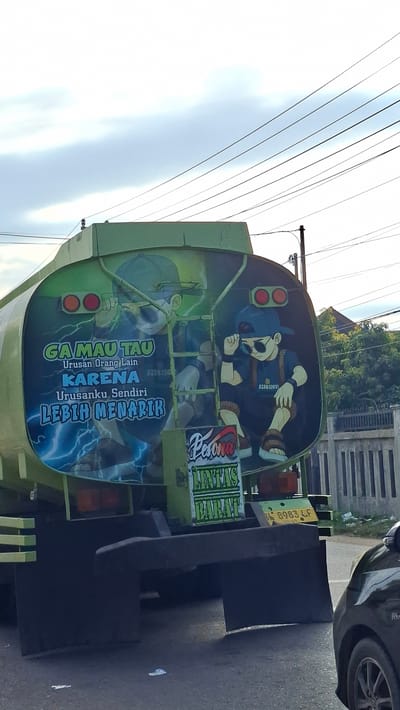 The image shows the rear of a green tanker truck. On the back of the tank, there is a large mural depicting two cartoon characters. The character on the left is looking through a spyglass, and the character on the right is adjusting their sunglasses. Text on the mural reads "GA MAU TAU Urusan Orang Lain KARENA Urusanku Sendiri LEBIH MENARIK" which translates to "DON'T CARE About Other People's Business BECAUSE My Own Business IS MORE INTERESTING." Below the mural, there is a smaller sign with the text "PESONA LINTAS BARAT" which translates to "CHARM OF WESTERN CROSSING." A license plate with the number "BL 8983 LF" is visible on the back of the truck. The truck is parked on a road, with a grey car partially visible on the right side of the image. The background shows trees, a fence, and overhead power lines. The scene appears to be in Keudebayu, Indonesia, as indicated by the license plate which starts with "BL," commonly used for vehicles registered in Aceh, Indonesia, and the overall environment.