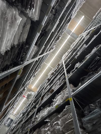 This is a downward-facing, wide-angle, interior shot of a warehouse in Holon, Israel, likely taken during daylight hours due to the bright overhead lighting. The scene is dominated by rows of tall, metal shelving units filled with items covered in clear plastic wrap. The camera's perspective is angled sharply, looking down a seemingly endless corridor of shelves.
The objects on the shelves are uniformly packaged, making it difficult to discern individual items. However, the plastic wrapping creates a reflective surface that captures the stark overhead fluorescent lighting. The shelving structure is a prominent feature, composed of grey metal beams and supports. Some of the shelves have yellow tags with writing on them, suggesting inventory management or labeling. One tag clearly shows the number "3". Another tag visible near the bottom of the frame has text that appears to be "XL".
The overall impression is one of organized, large-scale storage. There are no people visible in the frame, and the activity taking place is implied to be passive storage and organization of goods. The setting is industrial and functional, with a concrete ceiling and the uniform illumination typical of a commercial or industrial space. The image conveys a sense of scale and order within the warehouse.