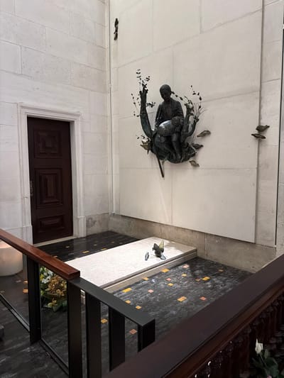 The image displays an interior view of the tomb of Francisco Marto, located within the Basilica of Our Lady of the Rosary of Fátima in Fátima, Portugal.
The scene is observed from a slight elevation, looking into a recessed area. A large, light-colored stone wall, composed of rectangular blocks, dominates the background. Centrally mounted on this wall is a bronze sculpture depicting a young boy, seated and holding a lamb. Stylized bronze branches with leaves extend from the sculpture, and additional bronze leaf motifs are affixed to the wall to the right.
Below the wall sculpture, a raised, rectangular slab of light-colored stone forms the tomb, positioned on a dark tiled floor. Two small bronze bird sculptures, one larger than the other, rest on this tomb slab. The floor surrounding the tomb features dark square tiles interspersed with smaller, scattered square tiles in shades of yellow, orange, and pink.
To the left, a dark brown wooden door with multiple panels and a light-colored frame is set into a wall of similar stone blocks. A dark brown wooden handrail and a black metal railing with vertical slats are prominent in the foreground, partially obscuring the lower left. Behind the railing on the far left, a white cylindrical object and yellow floral arrangements are partially visible. The lighting is even, suggesting an indoor, reverent environment.