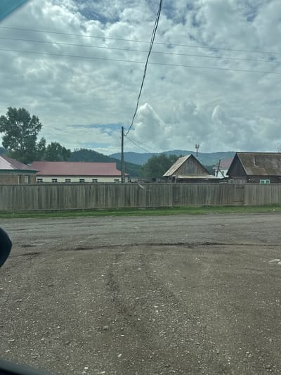 The image depicts a rural scene in the city of Yabogan, Russia. In the foreground, a gravel road with tire tracks is visible. A wooden fence runs horizontally across the mid-ground, separating the road from a row of houses and buildings behind it. The houses have various roofing materials, including red metal and corrugated metal. Some buildings have visible windows. Behind the houses, a line of trees and rolling hills can be seen, with mountains in the distance under a cloudy sky. Power lines are strung across the sky, supported by a wooden pole. The overall environment suggests a quiet, possibly remote, residential area.