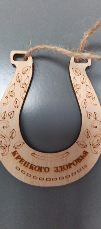 A wooden horseshoe charm hangs on a gray surface by a piece of twine. The charm is carved with decorative leaf and vine patterns on its upper arms. Along the bottom curve of the horseshoe, the Russian text "КРЕПКОГО ЗДОРОВЬЯ" (KREPKOVGO ZDOROVYA), which translates to "Good Health," is engraved, with a line of small, hollow squares beneath it. The horseshoe is positioned vertically, with its open end facing downwards. The twine loops through two small holes at the top of the horseshoe. The background is a plain, solid gray. There are no people or other objects visible in the image. This object is likely a souvenir or gift. The location of Tikhvin, Russia is not depicted in this image.