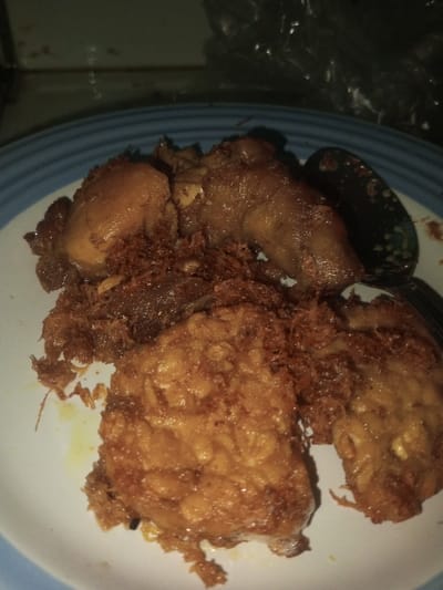 The scene, located in Karaikudi, India, displays a meal served on a white ceramic plate with a blue-banded rim. The plate contains multiple pieces of cooked poultry, likely chicken, characterized by a dark brown, somewhat glazed surface. These poultry pieces are mixed with and covered by a substantial amount of reddish-brown, finely shredded or crumbled, fried material. Additionally, two distinct, irregularly shaped, golden-brown fried patties are present; one large, mostly intact piece is in the lower-left quadrant of the plate, and a smaller, fractured piece is in the lower-right. A dark-colored serving spoon, possibly patterned, is partially visible on the right side, resting on the food. A metallic fork tine is also partially visible adjacent to the spoon. The lighting is dim, and the background is out of focus, showing a light-colored horizontal surface behind the plate and an indistinct, dark area above it. No individuals are present in the image.