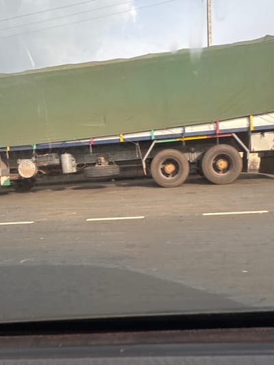 From an interior perspective, the scene depicts a portion of a moving truck, with its green canvas cover, tires, and undercarriage visible. The truck is traveling on a paved road with a painted white lane marker. Outside, the sky appears overcast. The location is Umuoye, Nigeria.