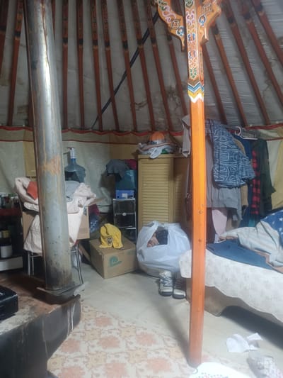 The image shows the interior of a traditional dwelling, likely a ger, in Ulaanbaatar, Mongolia. The structure features wooden poles supporting the roof, with decorative paint on some of the central supports. The walls are lined with fabric. The floor is covered with a patterned carpet.
The scene is cluttered with various objects, suggesting a lived-in space. In the foreground, a large metal pipe, possibly a chimney, stands to the left, attached to a dark, burnt-looking base that could be a stove. To the right of the pipe, there is a large white water cooler bottle.
Further into the dwelling, there are several stacks of boxes, some with labels, and various items of clothing and bedding are visible. A light-colored wooden cabinet with louvered doors is positioned against the wall. In front of this cabinet, a white plastic bag contains items, and next to it, a pair of grey and white hiking boots are placed on the floor.
On the right side of the image, a bed or sofa is covered with a light-colored textured fabric and has some bedding or blankets on it, including a blue one with white stars. There are also various items of clothing hanging on racks or clotheslines in the background. Scattered on the floor are pieces of white paper. The overall impression is one of a functional, somewhat disorganized living space.