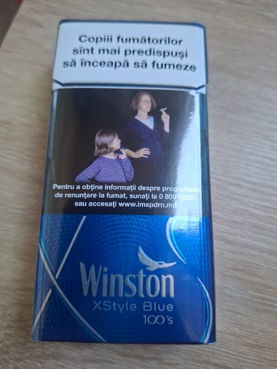The image shows a pack of Winston XStyle Blue 100's cigarettes, standing upright on a wooden surface. The cigarette pack has a blue color scheme with white text and a graphic of a bird. The top of the pack displays health warnings in Romanian. The text translates to: "Children of smokers are more predisposed to smoking" and "To obtain information about smoking cessation programs, call 0 800 10001 or visit www.imspdrn.md". The scene is likely indoors, given the lighting and the surface, and the image is a close-up, centered on the cigarette pack. There is no visible indication of the city of Durlești, Moldova.
