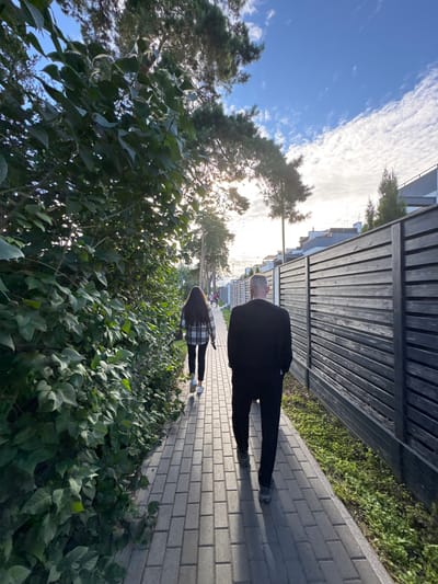 Two individuals, a man and a woman, are walking away from the camera on a paved sidewalk in Podstepki, Russia. The man is in the foreground, wearing a black suit jacket and pants. The woman is slightly ahead and to the left of the man, wearing a checkered shirt and dark pants. They are walking along a sidewalk bordered by lush green bushes on the left and a dark wooden fence on the right. In the background, there are modern houses and trees under a partly cloudy sky. The sun is shining, casting shadows on the sidewalk.