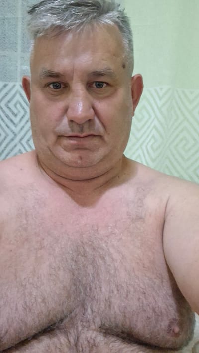 The image is a close-up, chest-up selfie of a middle-aged man. He has graying hair, a fair complexion, and is looking directly at the camera. His face is slightly flushed, and he has visible neck rolls and chest hair. He is shirtless. The background appears to be a bathroom with light green, patterned wallpaper. The man is located in Voinova, Moldova.