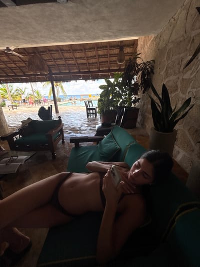 The image displays a semi-outdoor lounge area, likely part of a resort or hotel, located in Cozumel, Mexico. In the foreground, a female subject with dark hair is reclining on her left side on a teal-cushioned chaise lounge. She wears a dark two-piece bikini and holds a light-colored smartphone with both hands, viewing its screen. Her right arm is bent, supporting the device, while her left arm rests along her side. To the immediate left of the lounge, an open book with eyeglasses rests on a dark tiled floor next to a dark wooden armchair with a teal cushion.
The lounge area is covered by a thatched roof supported by wooden beams, from which two distinct light fixtures are suspended: one woven and one beaded. A stone-textured wall forms the right boundary of the covered space. Potted plants, including a tall Sansevieria, are positioned along this wall.
Beyond the covered area, an outdoor scene unfolds under a bright, partly cloudy sky. A swimming pool is visible in the midground, occupied by multiple individuals. Further in the background, the ocean is identifiable. Several palm trees are interspersed throughout the scene. Near the pool, outdoor tables and chairs are arranged, with other individuals seated. The ground surrounding the pool appears wet.