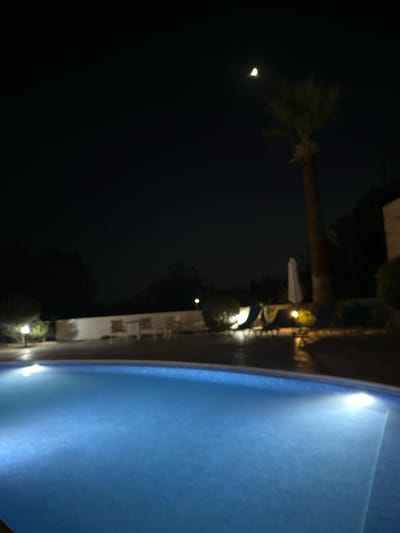 The image depicts a nighttime scene at a swimming pool in Peyia, Cyprus. A crescent moon is visible in the dark sky, with a palm tree to its right. The swimming pool, illuminated by underwater lights, occupies the foreground and mid-ground, appearing to be a resort or hotel property. In the background, a white wall with chairs and tables is visible, suggesting an outdoor seating area. There are lounge chairs and an umbrella near the pool. The overall impression is a peaceful, serene evening at a holiday destination.