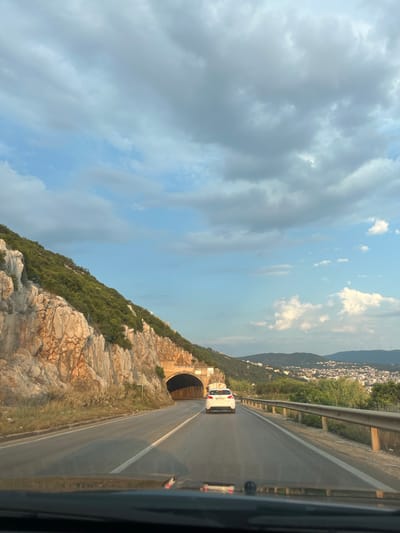 The image depicts a road curving towards a tunnel carved into a rocky mountainside, densely covered with green vegetation. A white car is driving towards the tunnel entrance. The road is asphalt, with a white line marking the lane. A metal guardrail runs alongside the road. The sky is a mix of blue and grey clouds, suggesting it might be late afternoon or early evening. In the distance, beyond the mountains, a town with white buildings is visible on the slopes. The scene appears to be a scenic route, possibly in a coastal or mountainous region. There is no visible text in the image. The image is taken from inside a vehicle, likely the passenger seat, given the perspective.