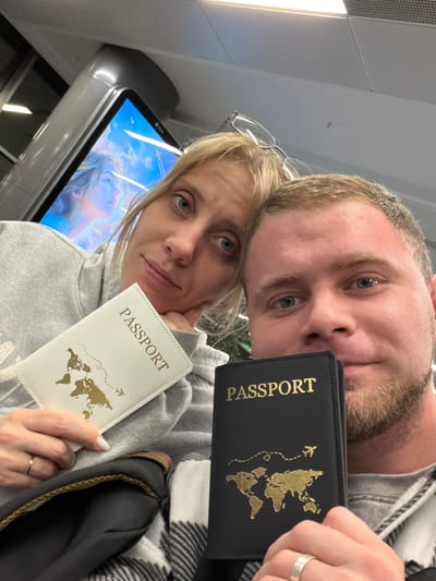 The scene depicts two individuals, one female and one male, captured in an indoor selfie-style photograph. The scene is an indoor setting in the city of Boldino, Russia.

The female, positioned on the left, has blonde hair, blue eyes, and wears glasses pushed onto the top of her head. She is wearing a grey hooded sweatshirt and holds a white passport cover in her left hand, featuring the word "PASSPORT" and a gold world map design. A thin silver ring with a small stone is visible on her left ring finger. Her chin rests on her right hand.

The male, positioned on the right and partially obscured by the female, has light brown hair, blue eyes, and light brown facial stubble. He wears a grey garment and a black and white plaid pattern, possibly a blanket or scarf, is draped over him. He holds a black passport cover in his right hand, which also displays "PASSPORT" and a gold world map design, complete with an airplane icon and a dashed flight path. A plain silver band is visible on his left ring finger.

In the background, a digital display screen shows a partial image of a person against a blue, cloudy sky. Overhead, a standard drop ceiling with recessed lighting fixtures is visible, along with a grey structural support column. A dark bag or object is partially visible near the female's left arm.