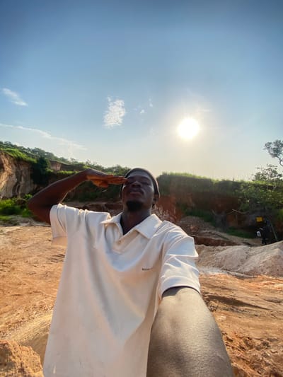 A young Black man, wearing a light-colored collared shirt and a dark beanie, salutes while standing in a dry, rocky, and dusty outdoor environment. Behind him, a natural landscape features hills with green vegetation and exposed reddish-brown earth. The sun is visible in the bright blue sky, casting light and shadows on the scene. The image appears to have been taken in a quarry or a similar geological site. The location is identified as Ufu Esioni, Nigeria.