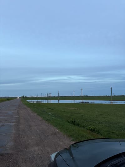 The image shows a straight, narrow dirt road in Azyakul', Russia. Power lines run along the roadside through a flooded, grassy field. A portion of a dark-colored car is visible in the right foreground. The sky is overcast with light blue tones. The landscape appears rural.
